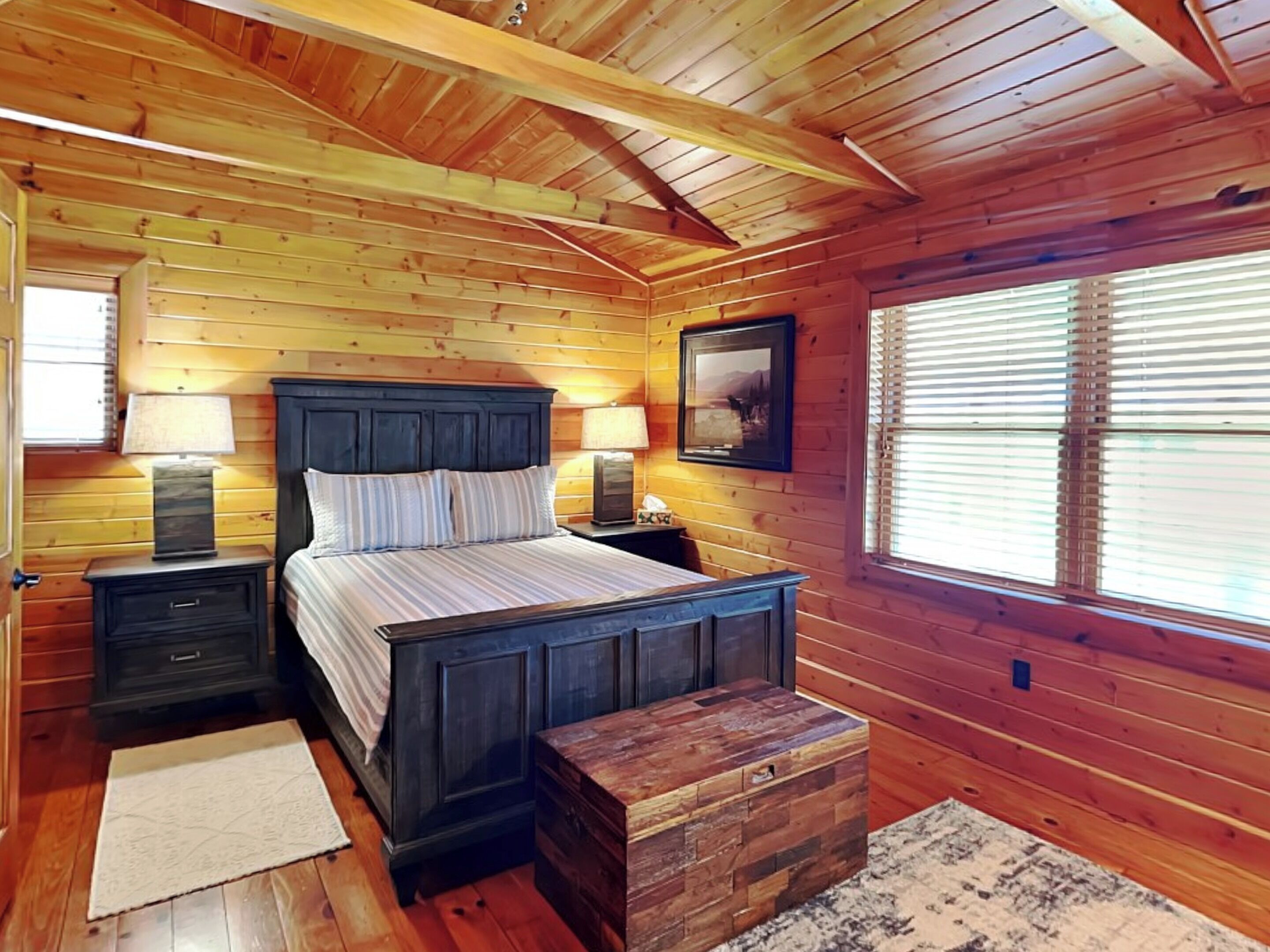 A wooden cabin bedroom with a bed, bedside tables, lamps, a window with blinds, and a wooden chest.