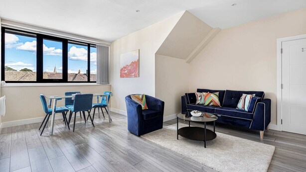 A spacious living room with a high ceiling, a dining area with blue chairs, a dark blue sofa with colorful cushions, and a large window offering a view of the sky and buildings. - City Service Lets - Large Cheam 1 bed with parking - Long stays welcome (London)