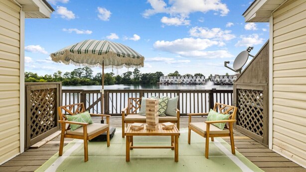 A deck with wooden furniture and a striped umbrella, overlooking a body of water and buildings.