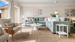 A living room with a sectional sofa, a round coffee table, and a small white table. There are two side tables with lamps, a potted plant, and artwork on the walls.