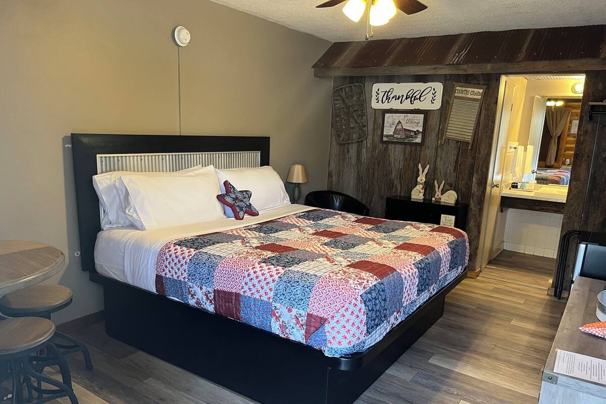 A bedroom with a large bed, a wooden headboard, a quilt, a nightstand, a chair, and a small table.