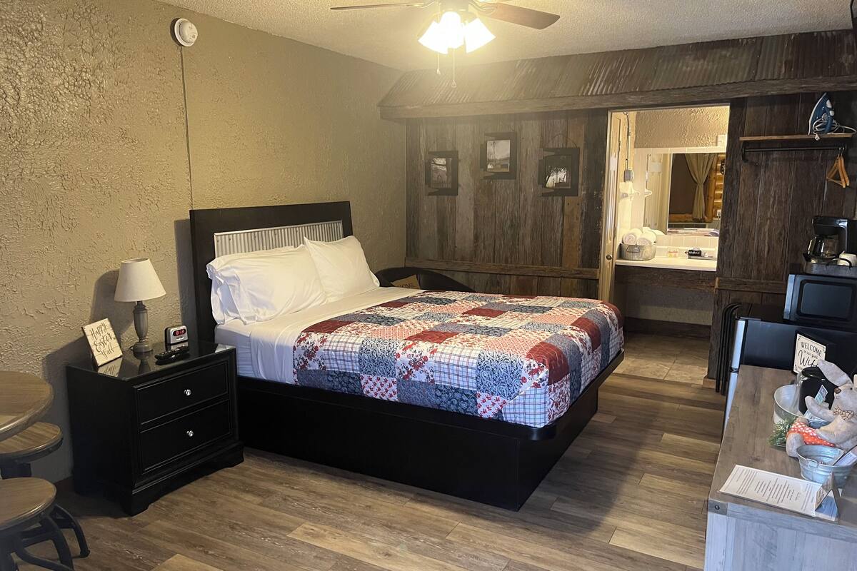 A bedroom with a bed, bedside tables, a nightstand, a desk, a chair, a ceiling fan, and a bathroom visible through an open door.
