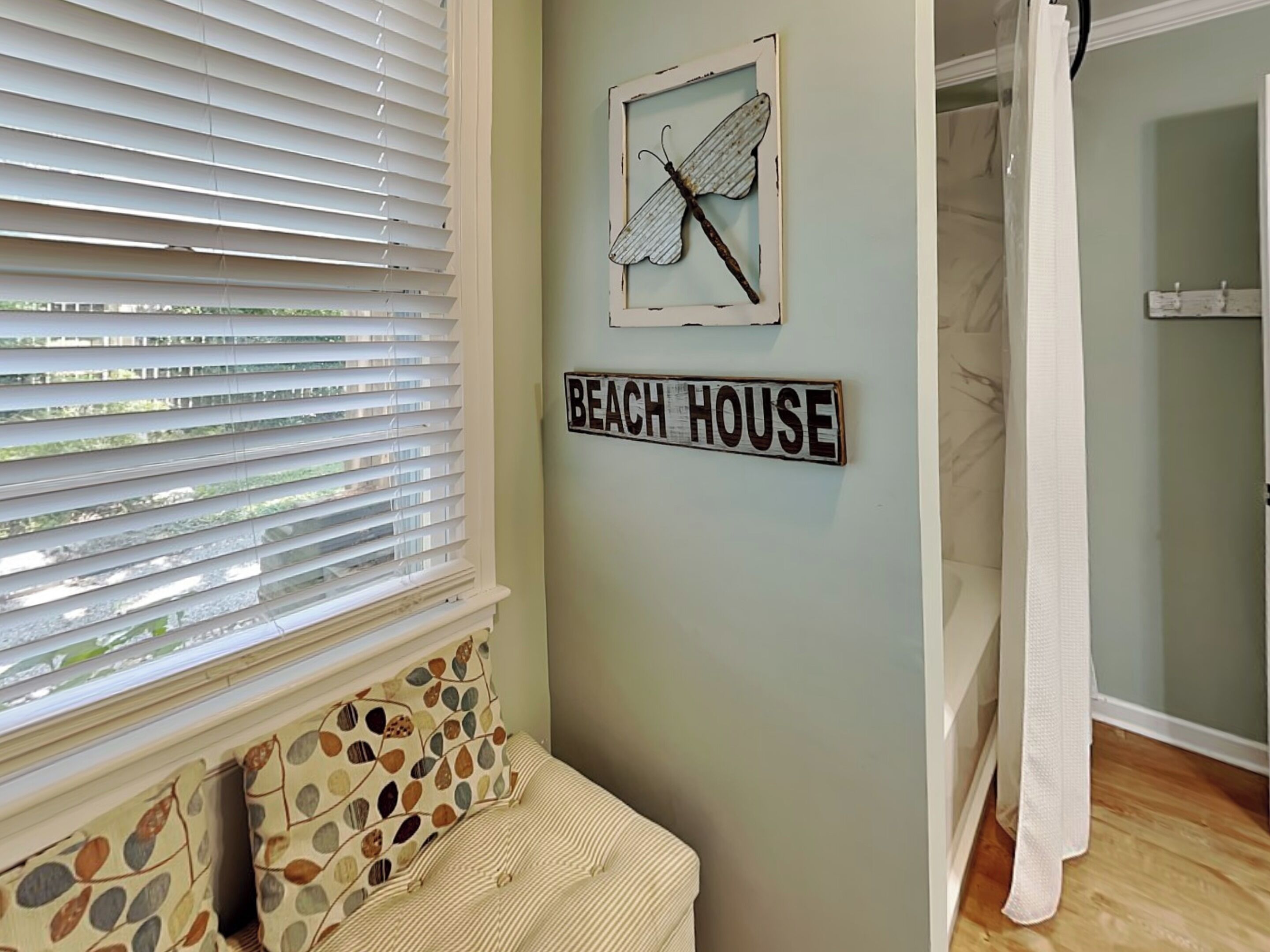 A room with a window with blinds, a wall-mounted sign reading "BEACH HOUSE", a framed artwork of a dragonfly, and a small bench with patterned cushions.