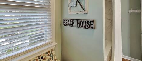 A room with a window with blinds, a wall-mounted sign reading "BEACH HOUSE", a framed artwork of a dragonfly, and a small bench with patterned cushions.