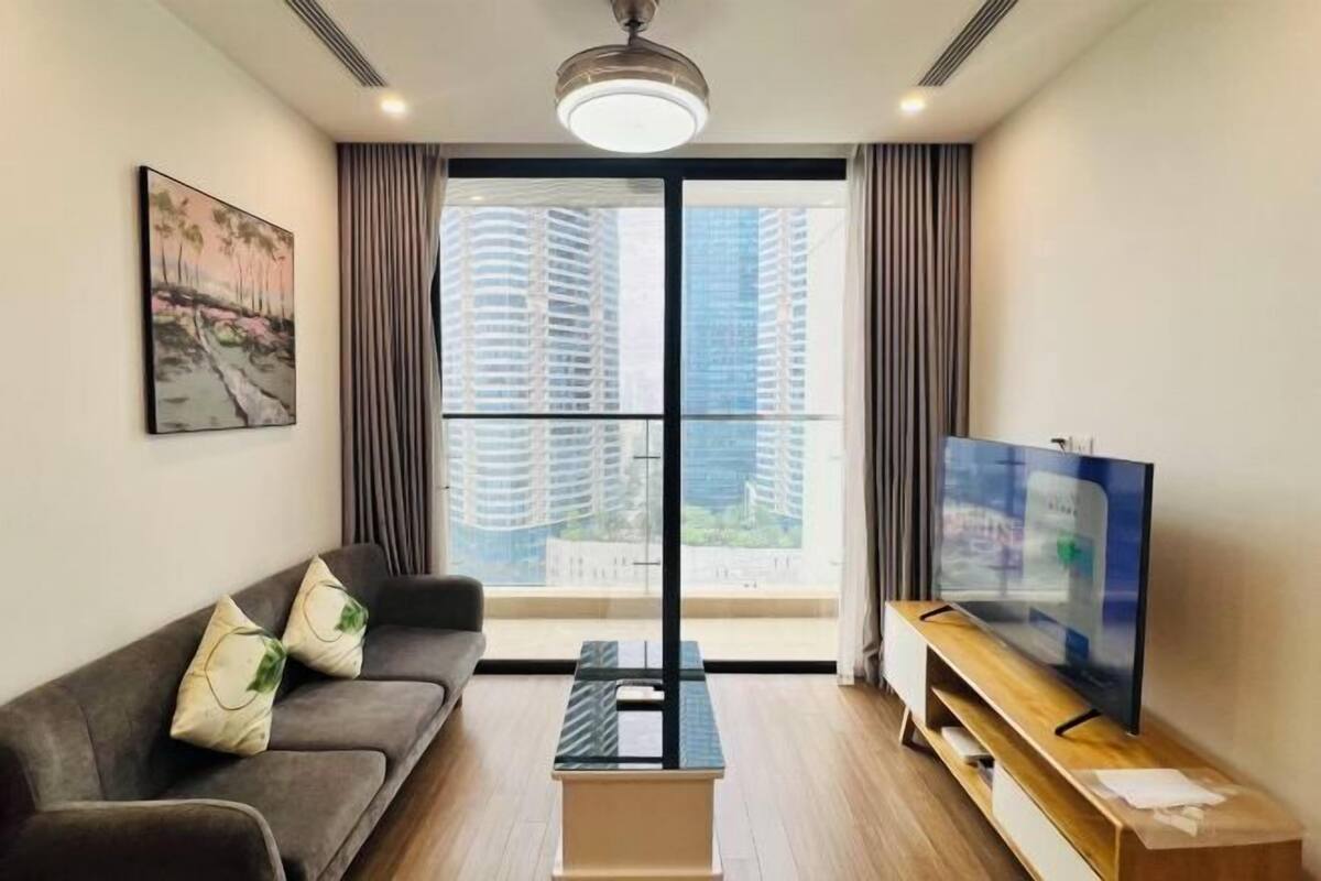 Uma sala de estar moderna com sofá, mesa de centro, TV e uma janela grande com vista para edifícios altos.