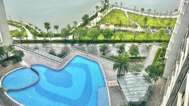 A swimming pool with a heart-shaped design, surrounded by a landscaped area with palm trees and a view of a hill with a fence. - Vinhomes Skylake Ha Noi Services Apartments - GalaxyHouse (Hanoi)