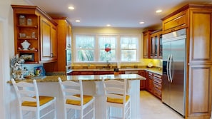 A kitchen with wooden cabinets, a granite countertop, a stainless steel refrigerator, and a window with a view of trees. - 6 Waterside Walk by Tybee Vacation Rentals (Tybee Island)