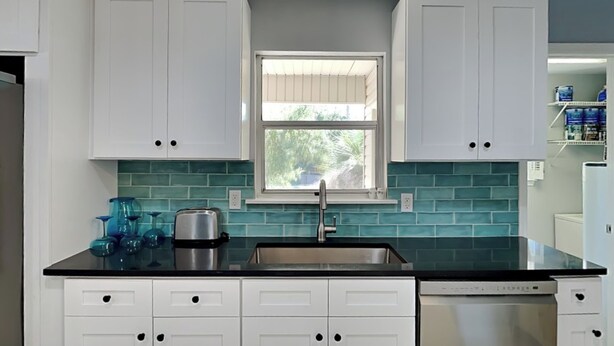 A kitchen with white cabinets, a black countertop, a stainless steel sink, and a window above the sink. - Lillypad by Tybee Vacation Rentals (Tybee Island)