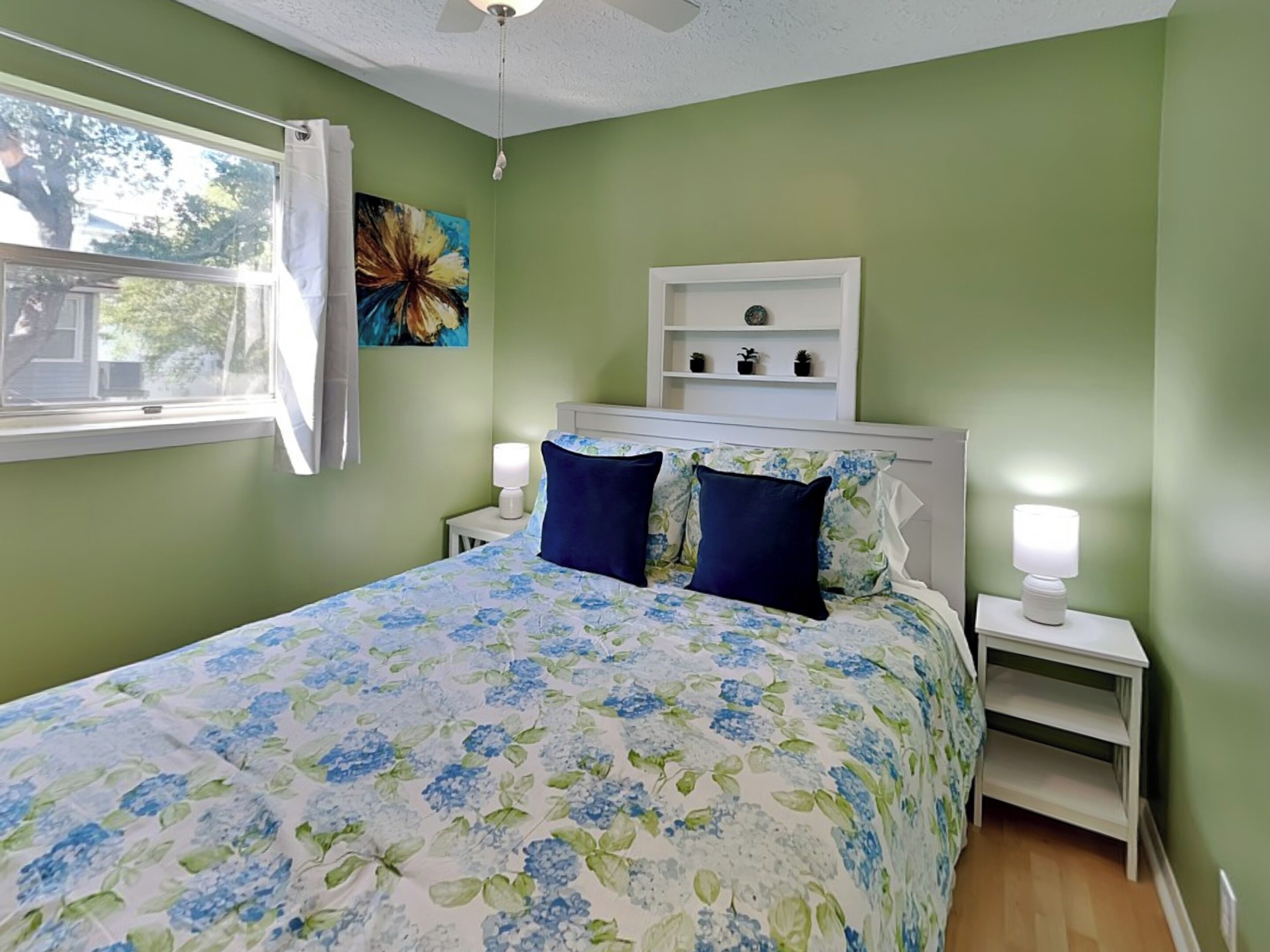 A bedroom with a floral bedspread, two pillows, a nightstand with a lamp, a shelf with decorative items, and a window with curtains.