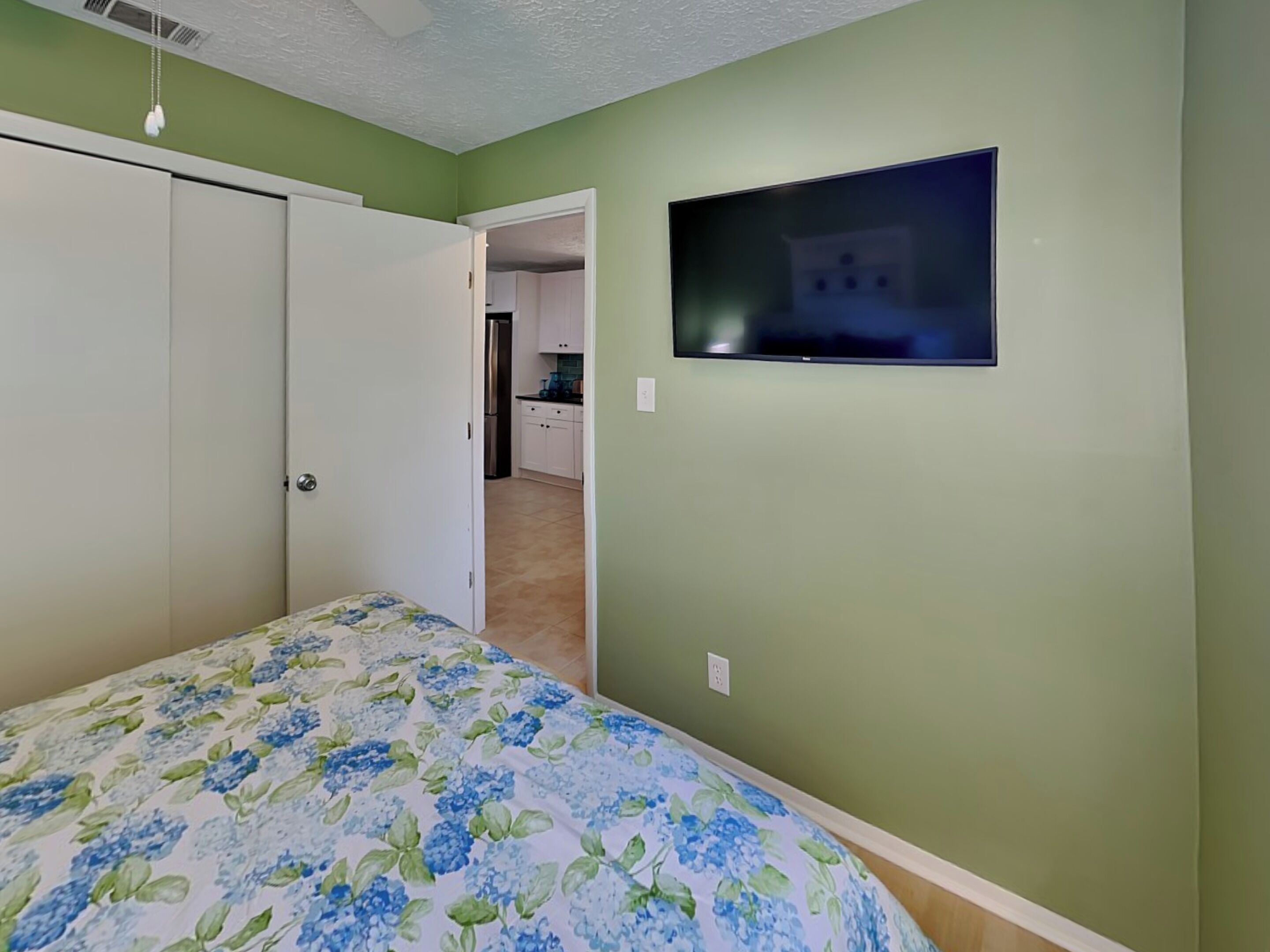 Un dormitorio con una cama, un televisor en la pared y una cocina al fondo.