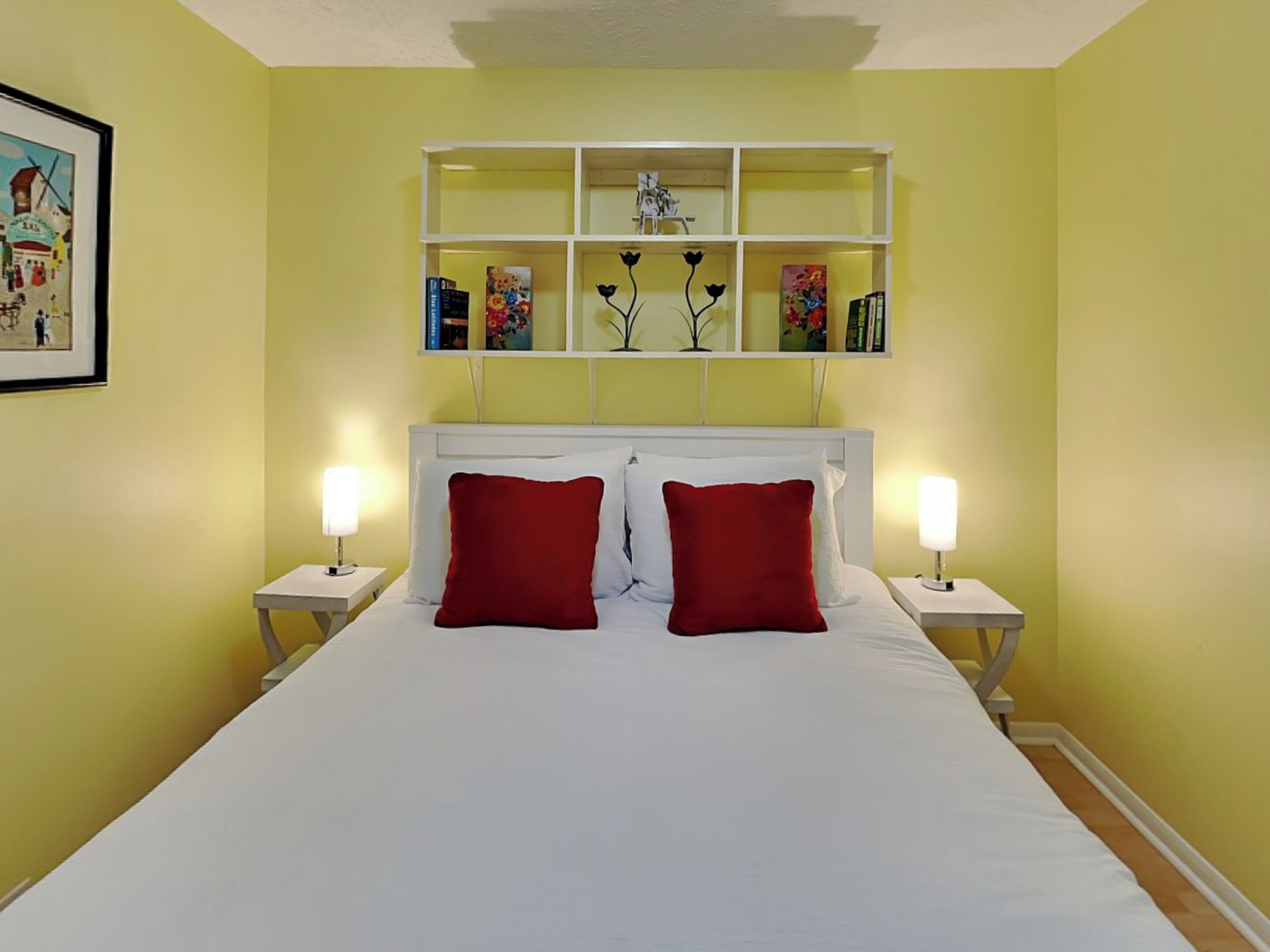 Una habitación de hotel con una cama grande, dos almohadas rojas, cabecera blanca, mesitas de noche con lámparas y un estante adosado a la pared con libros y objetos decorativos.