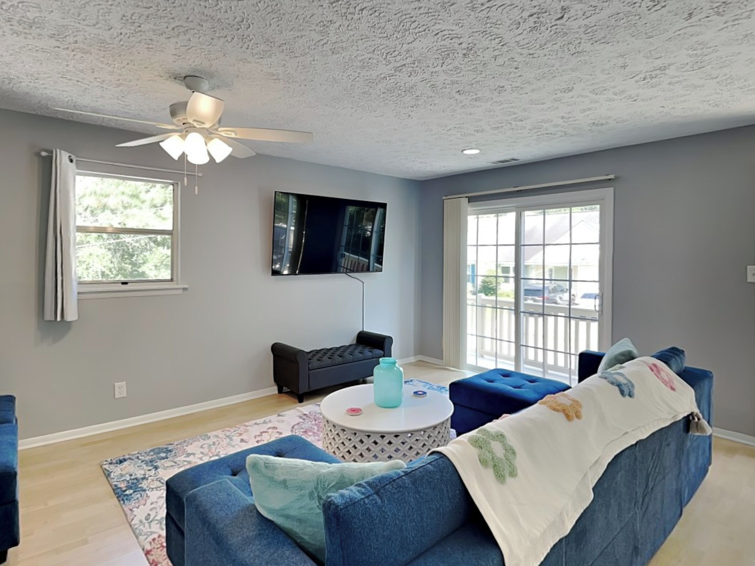 A modern living room with a blue sofa, a round coffee table, a ceiling fan, a flat-screen TV, and large windows.