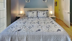 Un dormitorio con una cama, dos mesitas de noche, una mesita auxiliar, una puerta y una pared con dos cuadros enmarcados.