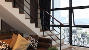 A modern interior with a staircase, a sofa with patterned cushions, and a large window with a city view. - Arte Mont Kiara By Yan Yee (Kuala Lumpur)