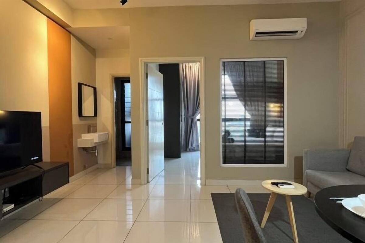 Een moderne woonkamer met een flatscreen televisie, een grijze bank, een klein rond tafeltje en een raam met zonwering.