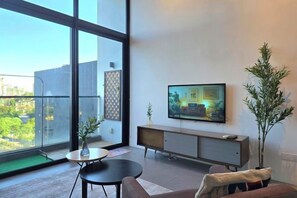 A modern living room with a large window, a flat-screen TV mounted on the wall, a sofa, a coffee table, and a small side table. - The Establishment Menara Teguh Bangsar Menara UOA KL Sentral (Kuala Lumpur)