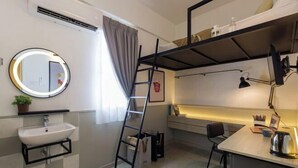 A compact room with a bunk bed, a bedside table with a lamp, a small desk, a round mirror, a sink, and a window with curtains. - Upstairs Downstairs CoLiving Bukit Bintang (Kuala Lumpur)