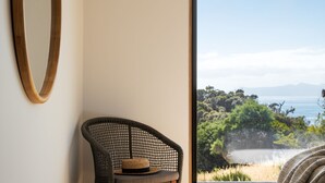 A room with a wicker chair, a wooden frame on the wall, and a view of a coastal landscape. - Rocky Ridge Itas - Hosted by L'Abode (Rocky Hills)