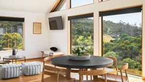 A modern room with a large window offering a scenic view of a forested area. There is a round table with chairs, a small white desk, and a black speaker. - Rocky Ridge Itas - Hosted by L'Abode (Rocky Hills)