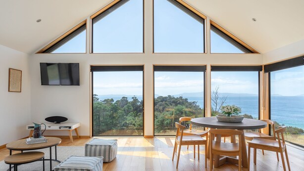 A spacious room with a high ceiling, large windows offering a view of the sea, a round dining table with chairs, and a flat-screen TV mounted on the wall. - Rocky Ridge Itas - Hosted by L'Abode (Rocky Hills)
