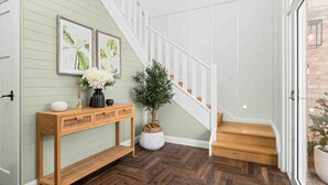 A wooden console table with a vase of flowers, a potted plant, and a wall-mounted picture frame in a hallway with herringbone-patterned flooring and a white staircase. - 4BDR, Fireplace, Pool & Pet friendly (Milton)