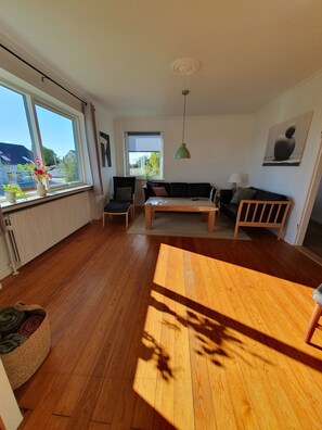 A spacious living area with a wooden floor, a sofa, a dining table, and large windows offering a view of the outside. - Lovely holiday home with views of fields (Sydals)