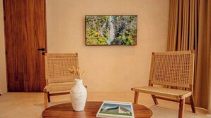 Um quarto com duas cadeiras de vime, uma mesa redonda de madeira, um vaso branco e um quadro na parede.