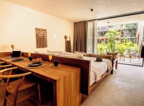 A modern hotel room with a wooden dining table, a sofa, and a large sliding glass door leading to a balcony with greenery. - Deluxe studio in Veleta w/private pool (Tulum)