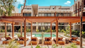 Area piscina moderna all'aperto con lettini in legno e una zona relax coperta, circondata da vegetazione rigogliosa e un edificio a più piani sullo sfondo.