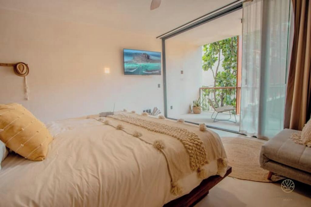 A bedroom with a bed, a sofa, a painting, and a sliding glass door leading to an outdoor area with a chair and a plant.