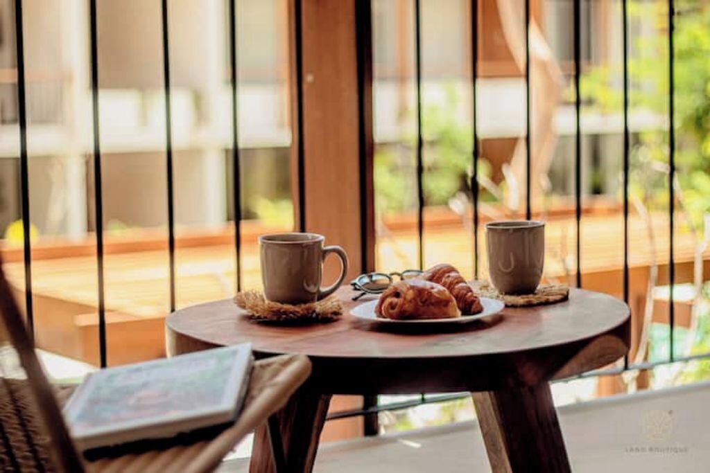 A round wooden table with two mugs and a plate of croissants, set on a balcony with a view of greenery.