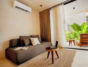 A modern living room with a sofa, a laptop on a small wooden stool, and a large window with curtains.