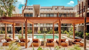 Uma área de piscina moderna ao ar livre, com espreguiçadeiras de madeira e um espaço de estar coberto, cercada por vegetação exuberante e um edifício de vários andares ao fundo.