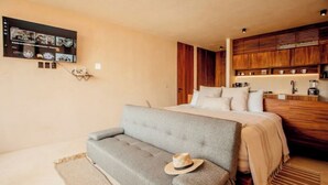Quarto moderno com sofá cinzento, cama com roupa de cama branca, armários de madeira e televisão na parede.