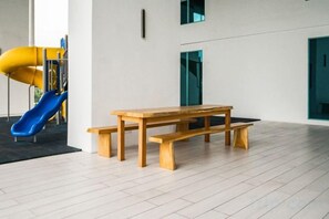 A wooden table and benches in a room with a white floor and a playground with a blue slide in the background. - Liwan, Baysuite by Ayuhouz (Kota Kinabalu)