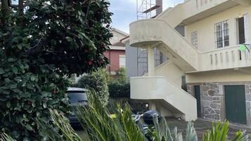 Un edificio de varios pisos con una escalera central, un coche estacionado en la entrada y un árbol con bayas rojas.