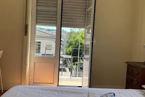 Una stanza con un letto, una cassettiera in legno e una porta scorrevole in vetro che dà su uno spazio esterno con vista su edifici e alberi.