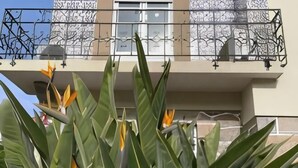 Ein Balkon mit schmiedeeisernem Geländer, eine Gebäudefassade und üppiges Grün im Vordergrund.