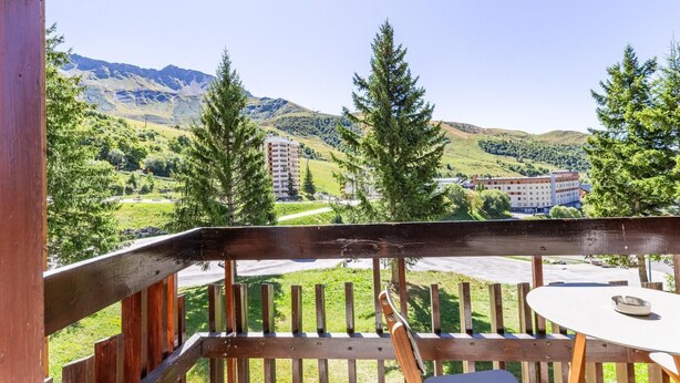 Une terrasse en bois avec une table et des chaises, d’où l’on peut admirer un paysage montagneux avec des bâtiments en arrière-plan.