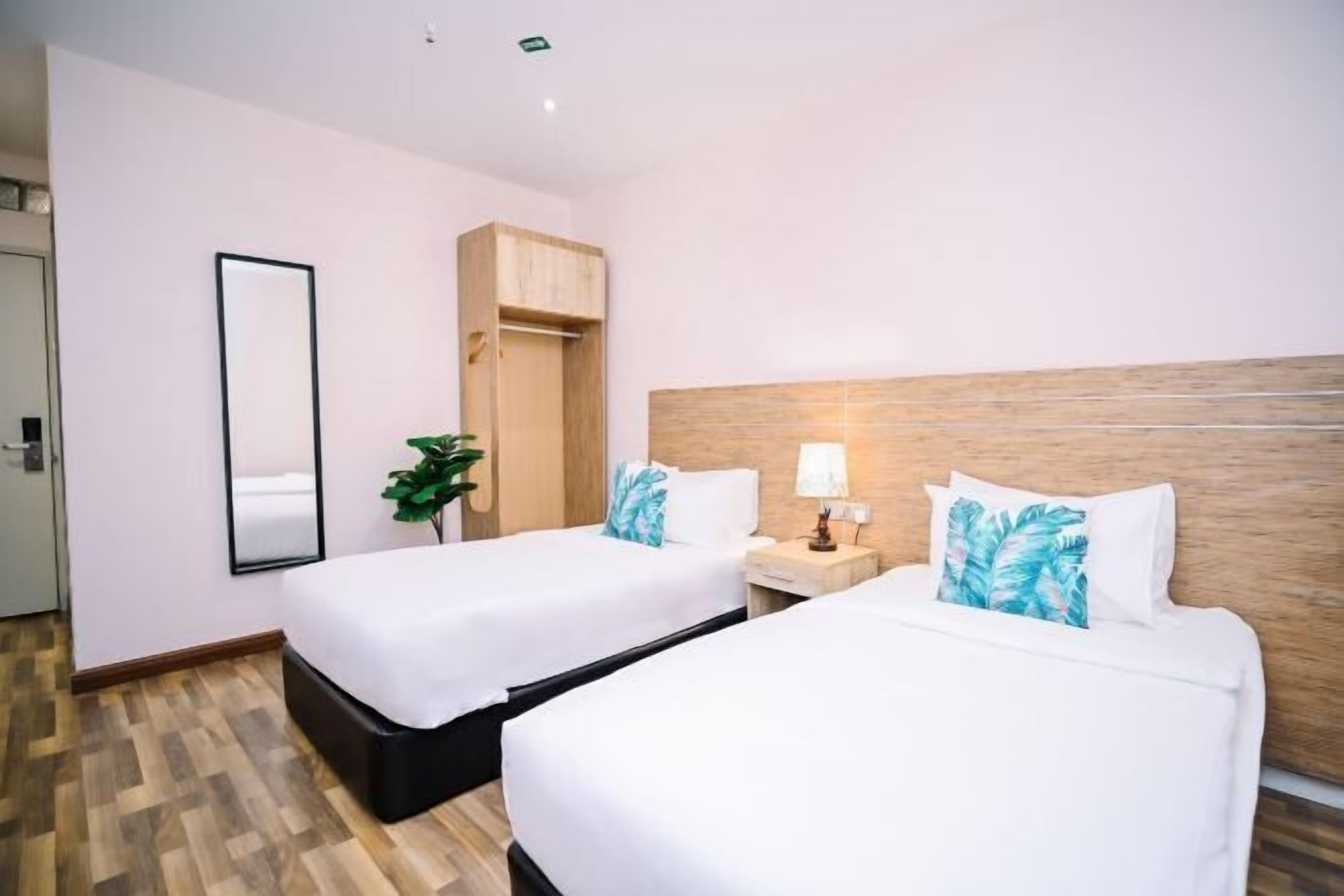 A hotel room with two beds, a wooden headboard, a nightstand with a lamp, a mirror, and a plant.