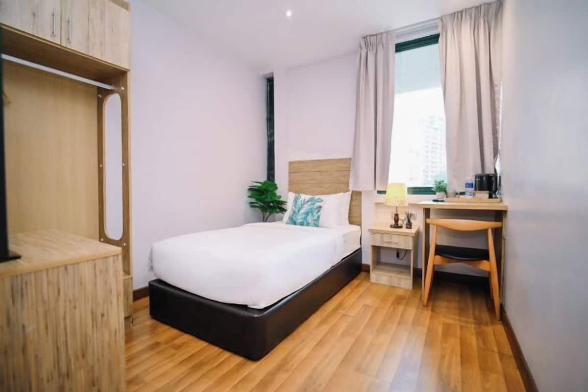 A hotel room with a bed, a desk, a chair, a plant, and a window with curtains.