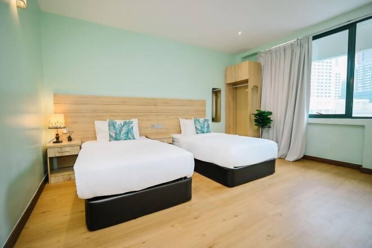 A hotel room with two beds, a nightstand, a mirror, a window with curtains, and a view of buildings.