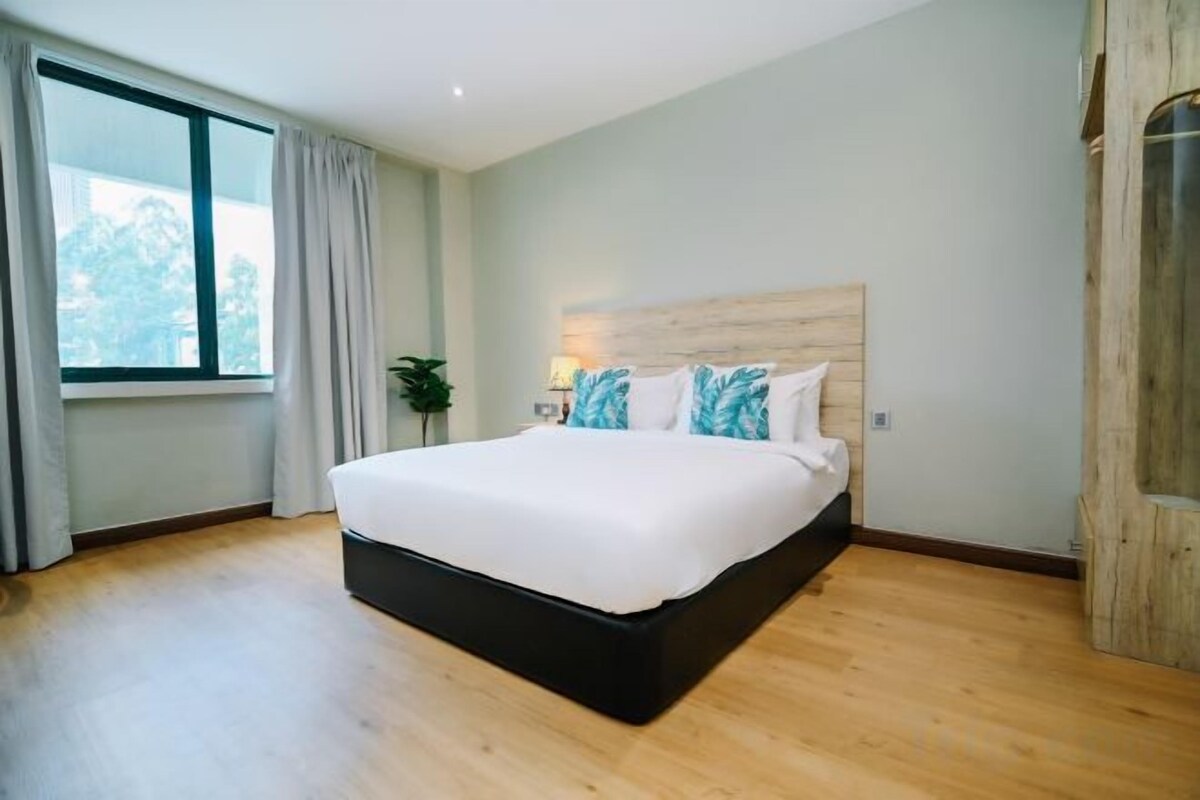 A modern bedroom with a large bed, a wooden headboard, a window with curtains, and a plant on the nightstand.