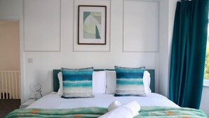 Un lit bien fait, recouvert d’une couverture verte et blanche, avec deux oreillers à motifs bleus et blancs, un tableau encadré au mur et une fenêtre ornée de rideaux turquoise.