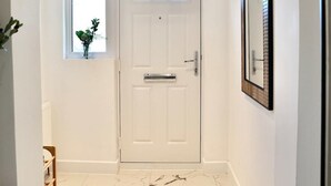 Un couloir avec une porte blanche, une petite fenêtre ornée d’une plante et un miroir accroché au mur.