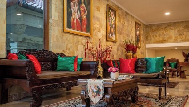 A traditional-style living room with ornate furniture, a coffee table, and a large window overlooking a building with a tiled roof. - Grand Melka Hotel (Bali)