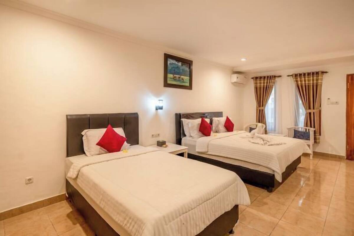 A hotel room with two beds, a nightstand, a wall-mounted lamp, a painting, and a window with curtains.