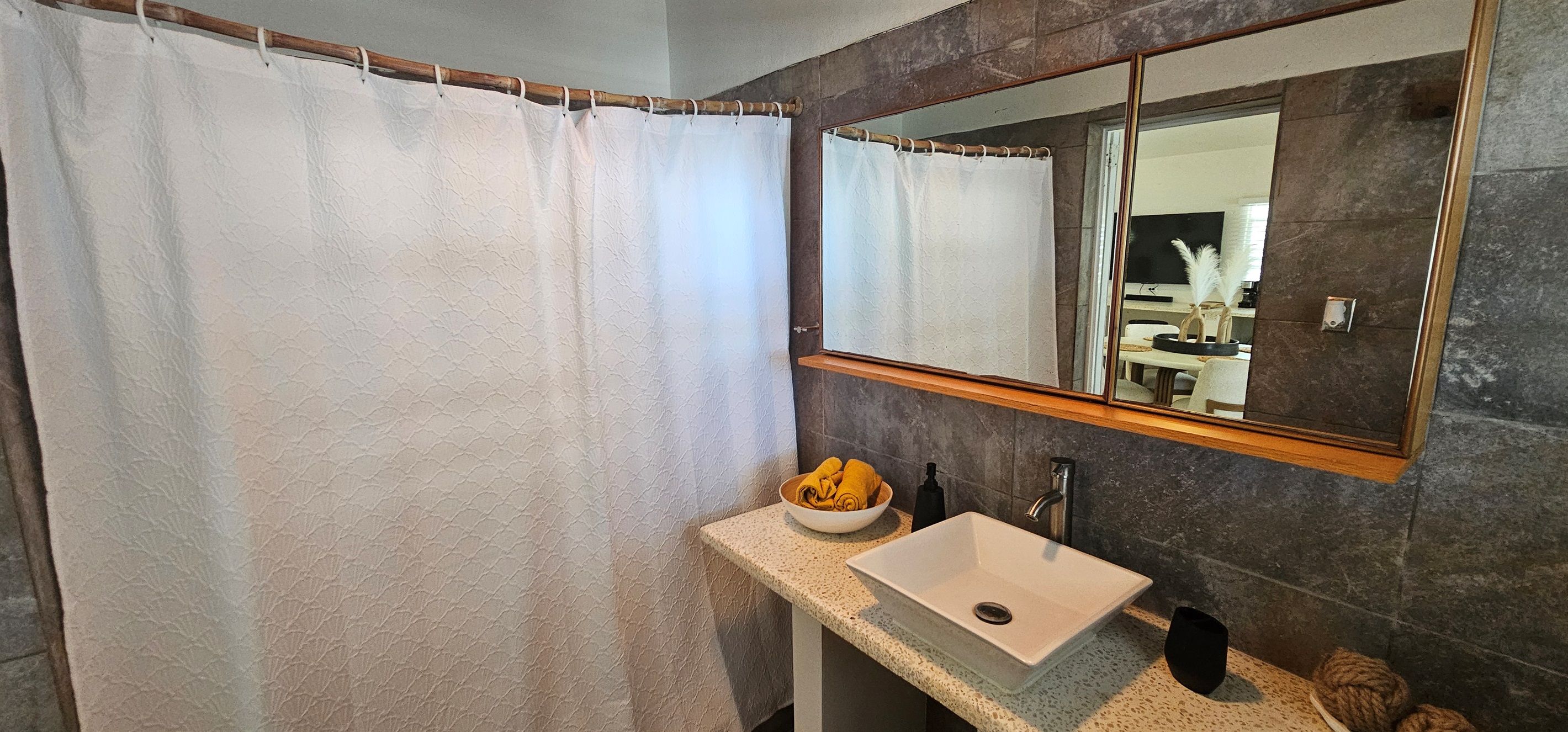 Un baño con cortina de ducha, un lavabo con un bol de toallas amarillas, un espejo y una botella de jabón.