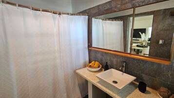 Un baño con cortina de ducha, un lavabo con un bol de toallas amarillas, un espejo y una botella de jabón.