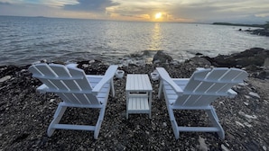 Två vita Adirondack-stolar placerade framför en stenstrand, med en solnedgång över vattnet i bakgrunden.